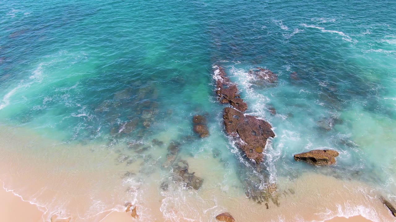 Aerial frame at Buzzards Beach with shallow blue-green water washing around a long central rock and a soft foam line touching the sandy beach, no visible people.
