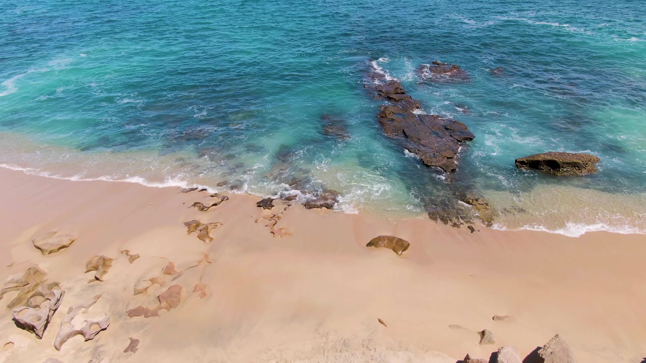 Aerial view of turquoise ocean waves meeting sandy beach and dark rocks.