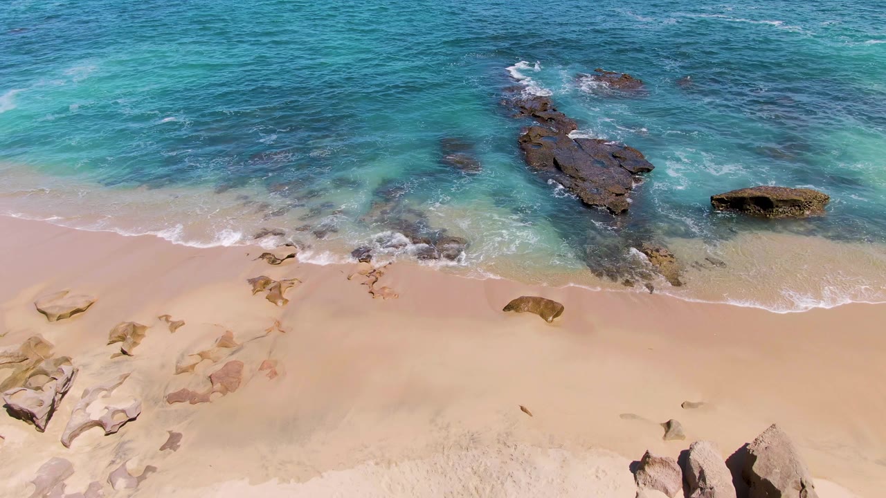 At Buzzards Beach in Baja California Sur, a thin wave line advances over pale sand while submerged and exposed rocks sit in clear turquoise water, with no visible people.