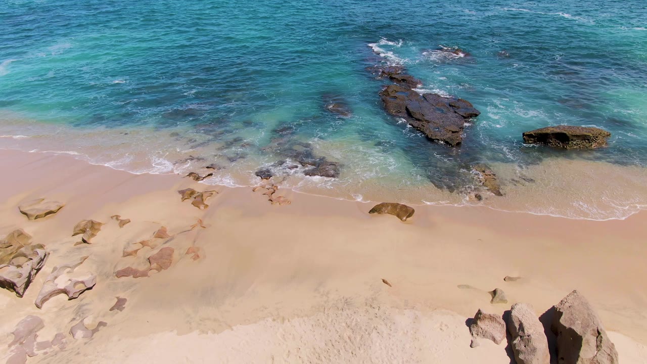 At Buzzards Beach in Baja California Sur, blue-green water spreads around clustered dark rocks above a smooth sandy beach, with no visible people.
