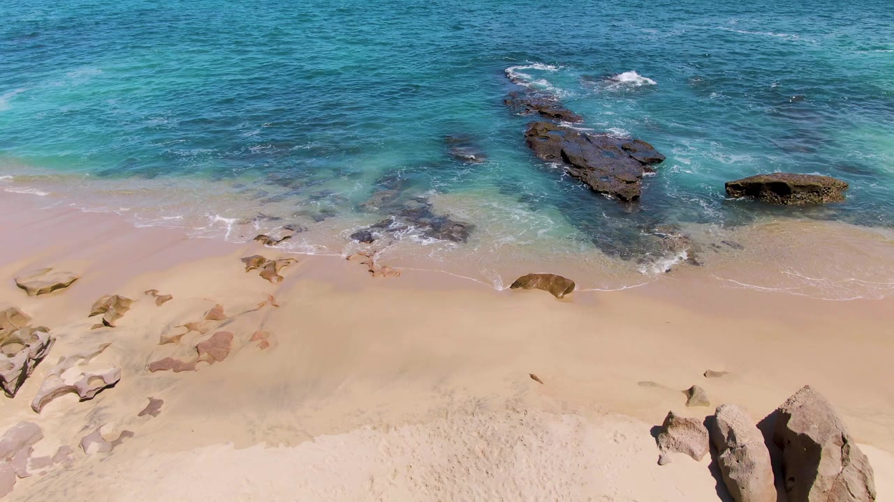 At Buzzards Beach in Baja California Sur, white foam breaks around dark rocks while clear water fades into deeper blue offshore, with no visible people.