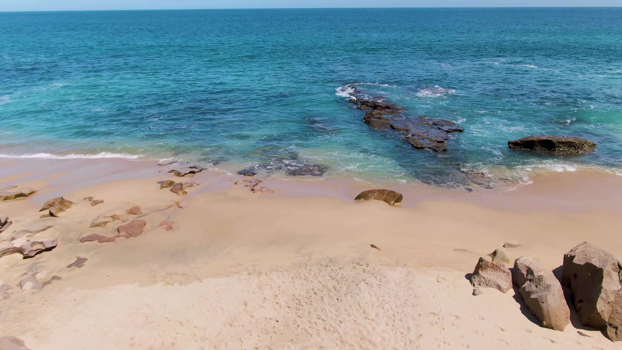 Wide aerial frame at Buzzards Beach, Baja California Sur, showing pale sand below turquoise water and dark rocky outcrops offshore with no visible people.
