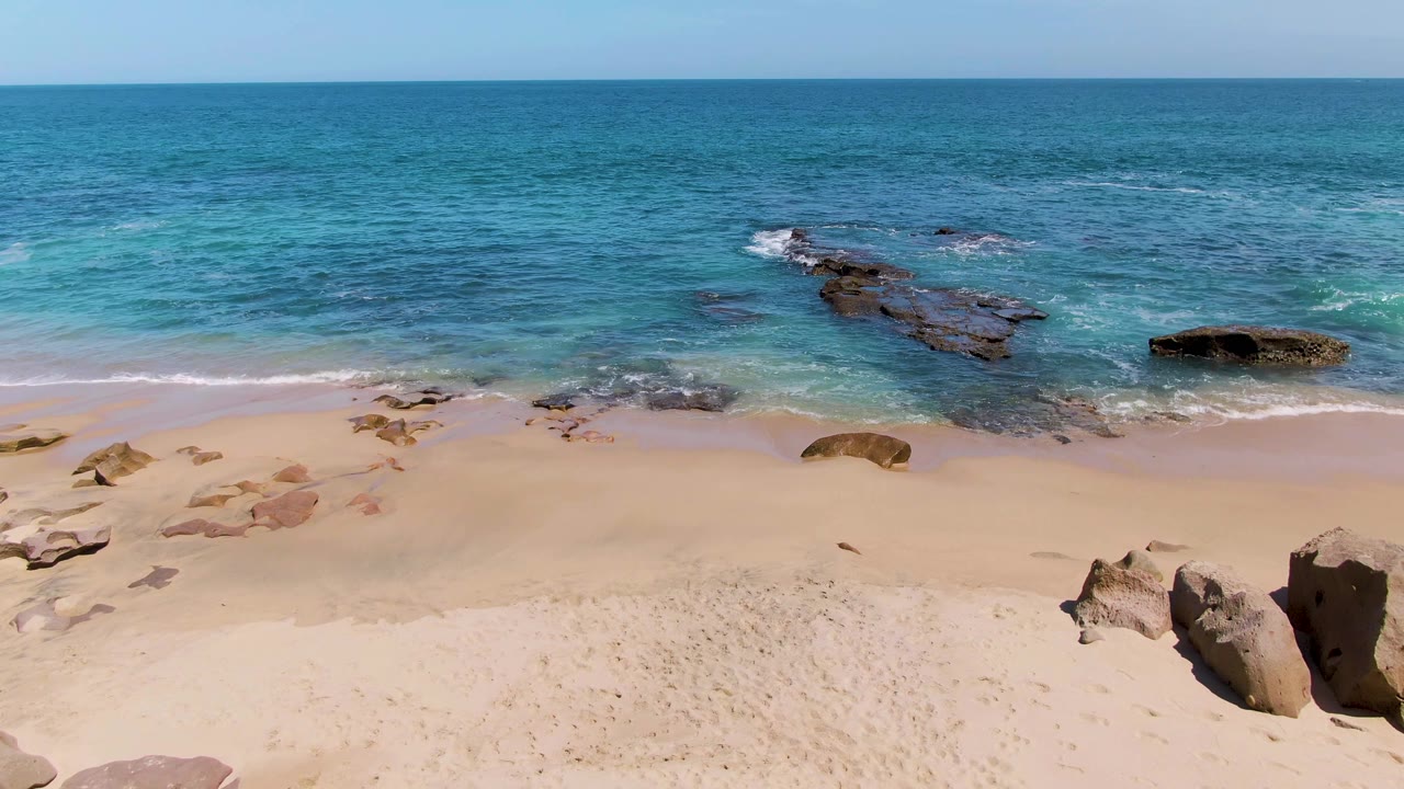 Aerial view over Buzzards Beach with the sandy foreground, blue-green water, scattered shoreline rocks, and no visible people.