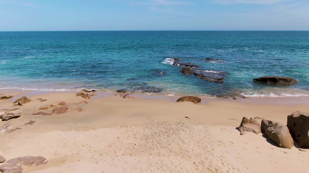 Buzzards Beach frame with turquoise surf around dark rock shelves, pale sand in the foreground, open ocean behind, and no visible people.