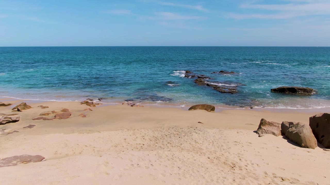 Empty sandy beach with low waves and offshore rocks under a blue sky