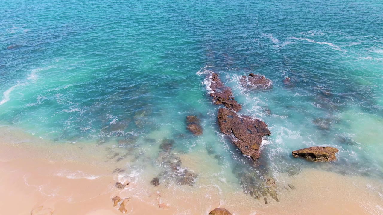 Aerial frame over Buzzards Beach in Baja California Sur, with turquoise water pushing white foam around dark rocks and no visible people.