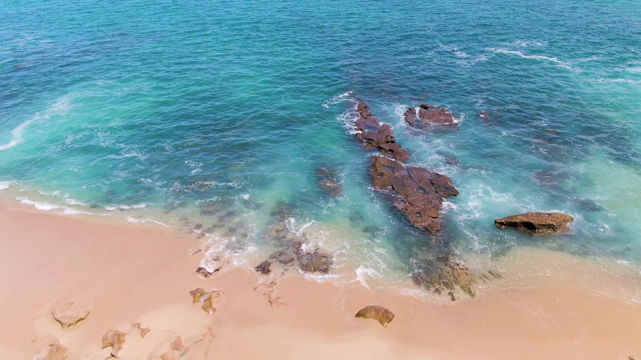 Aerial view at Buzzards Beach with exposed brown rocks near the center, clearer blue water above, pale sand below, and no visible people.