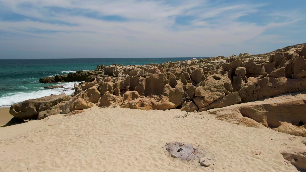 Opening view at Buzzards Beach, Baja California Sur, with pale sand in front, tan coastal rocks across the middle, turquoise ocean at left, and no visible people.
