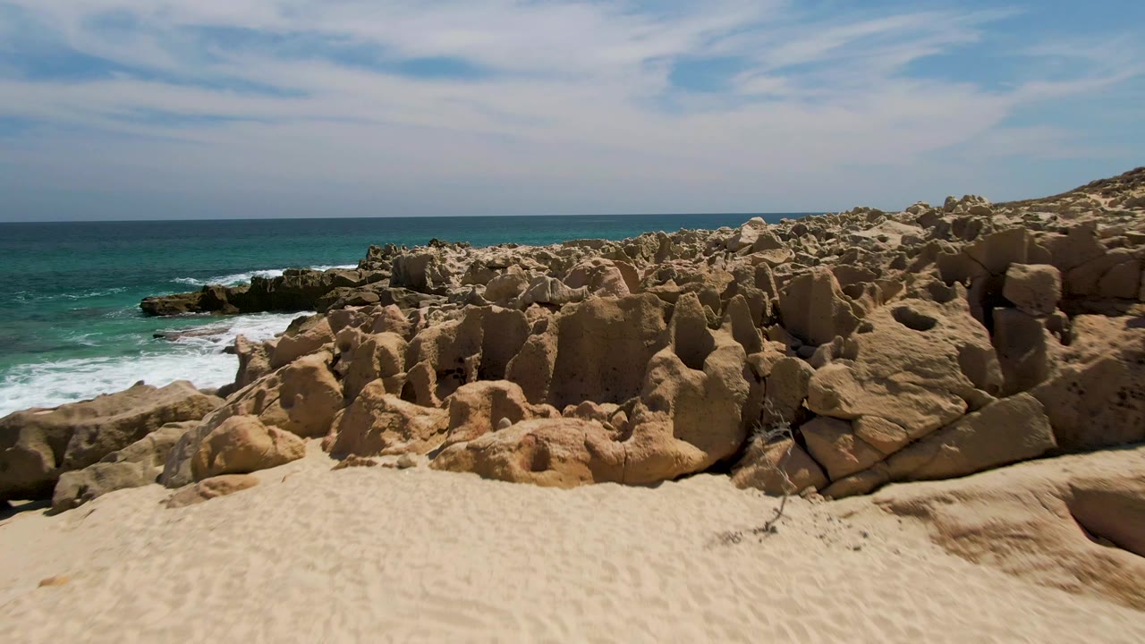 Mid-approach frame at Buzzards Beach, Baja California Sur, with textured sand below, jagged tan rock piles ahead, blue-green ocean to the left, and no visible people.