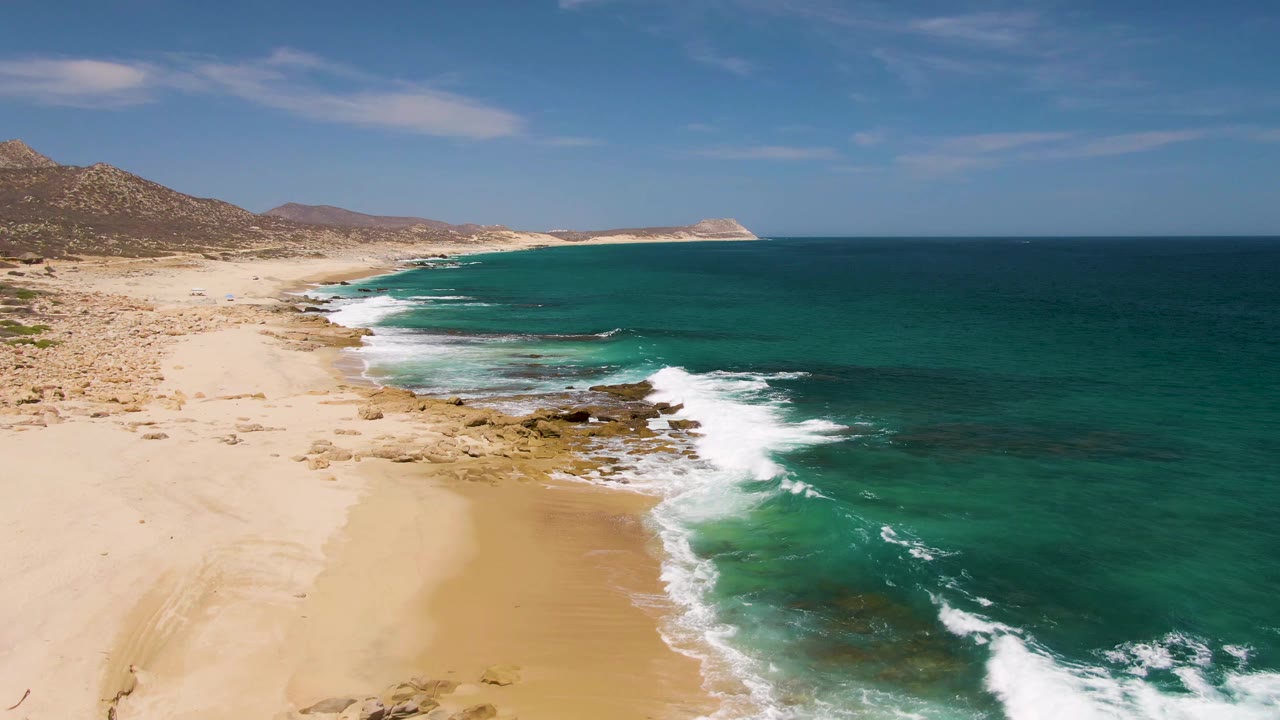Aerial view of sandy beach, turquoise surf, rocks, and distant hills.