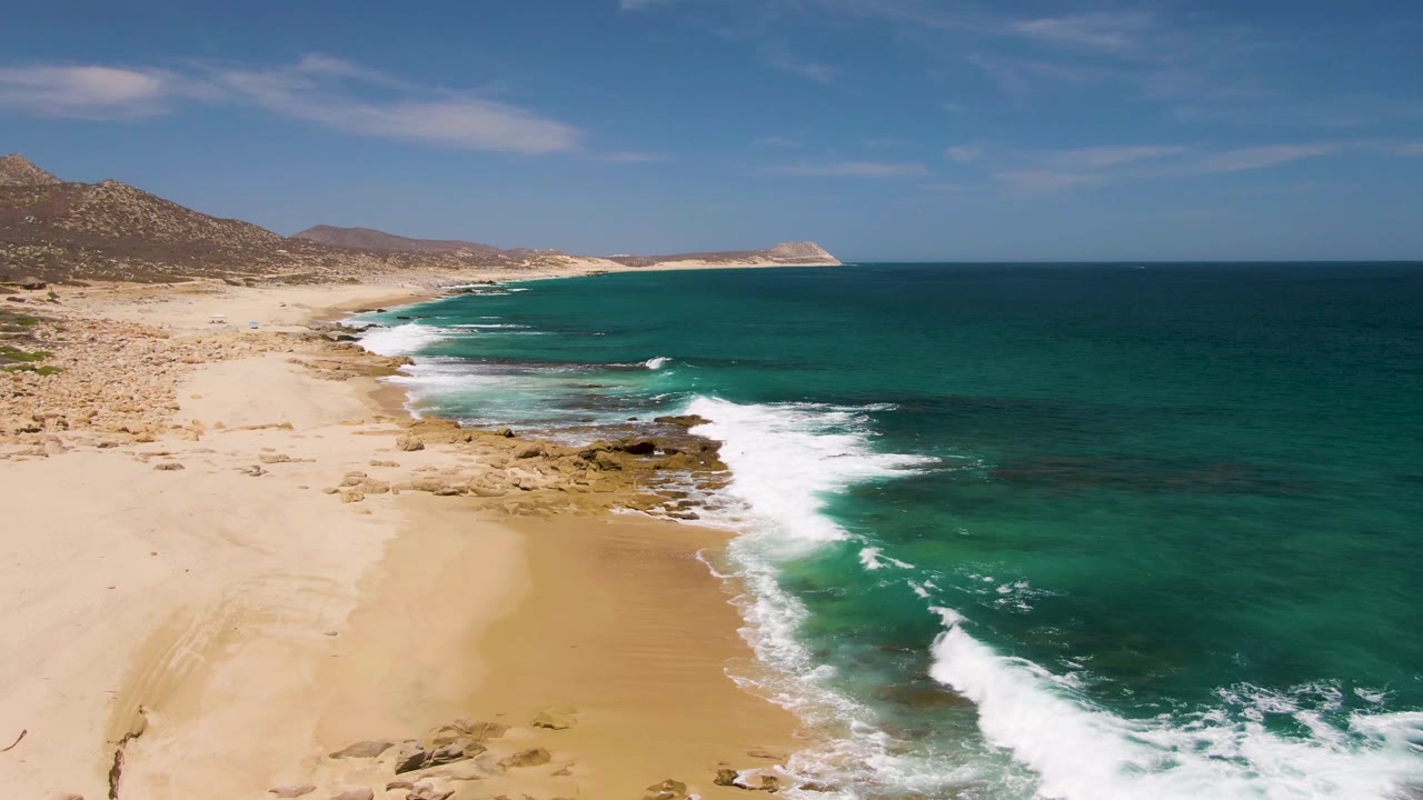 Second frame at Buzzards Beach shows white foam advancing along the sand, dark rock shelves near shore, green-blue water, distant hills, and no visible people.