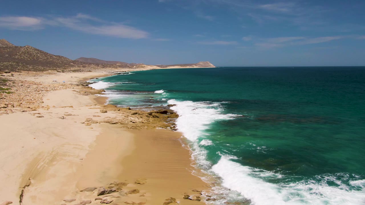 Aerial frame over Buzzards Beach with a broad band of white surf curling along the beach, exposed rocks near the waterline, open ocean, hills, and no visible people.