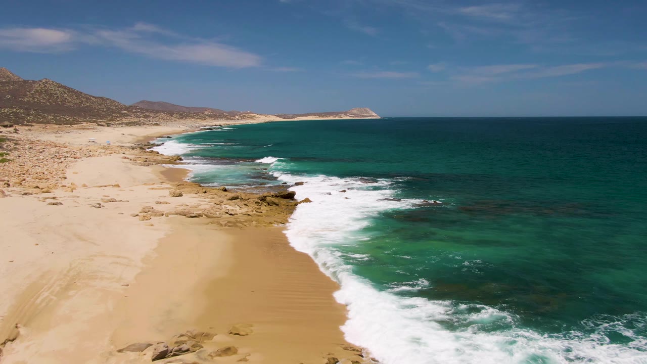 Mid-clip frame at Buzzards Beach showing white water spreading across the lower shoreline, turquoise shallows, tan sand, rocky points, hills, and no visible people.