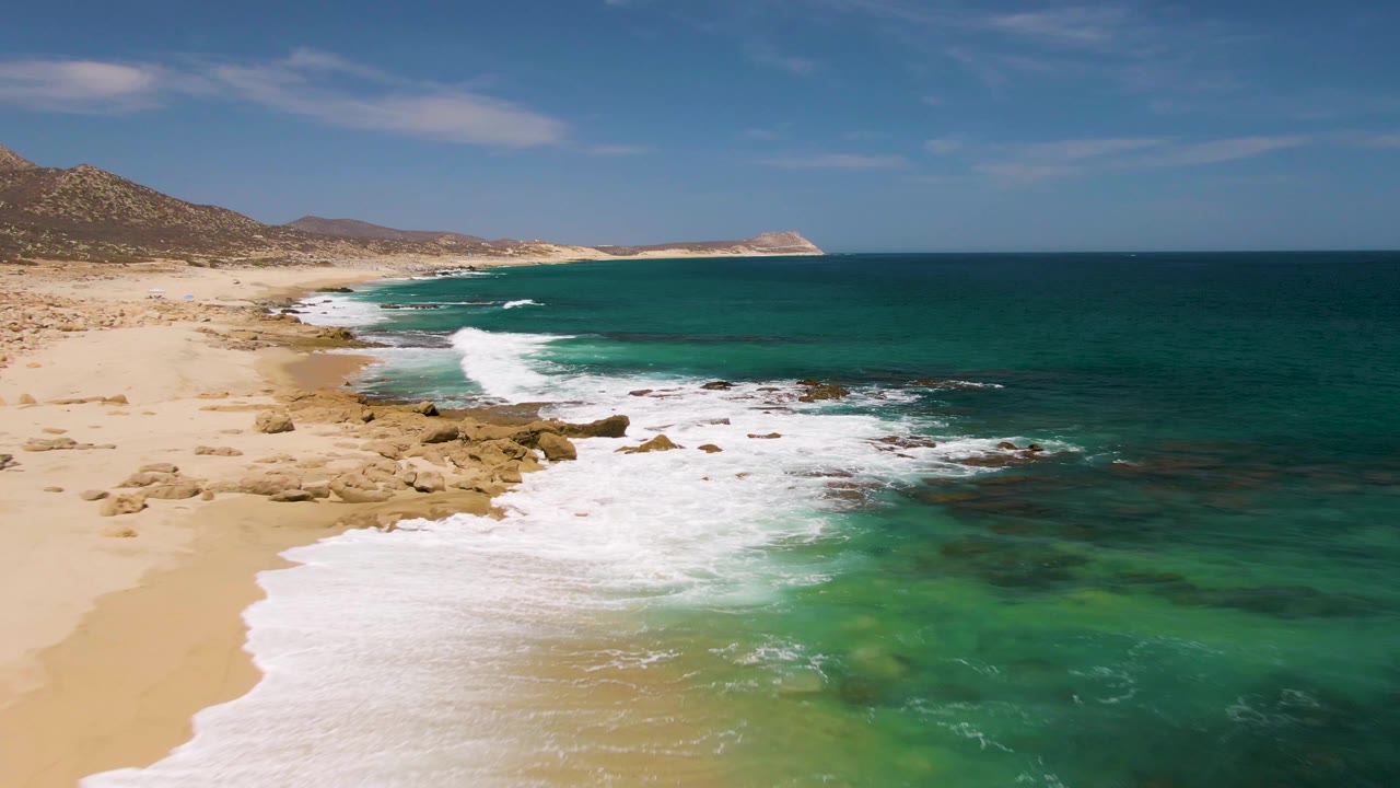 At Buzzards Beach in Baja California Sur, an aerial frame shows white surf running onto tan sand beside scattered rocks, with turquoise water and no visible people.