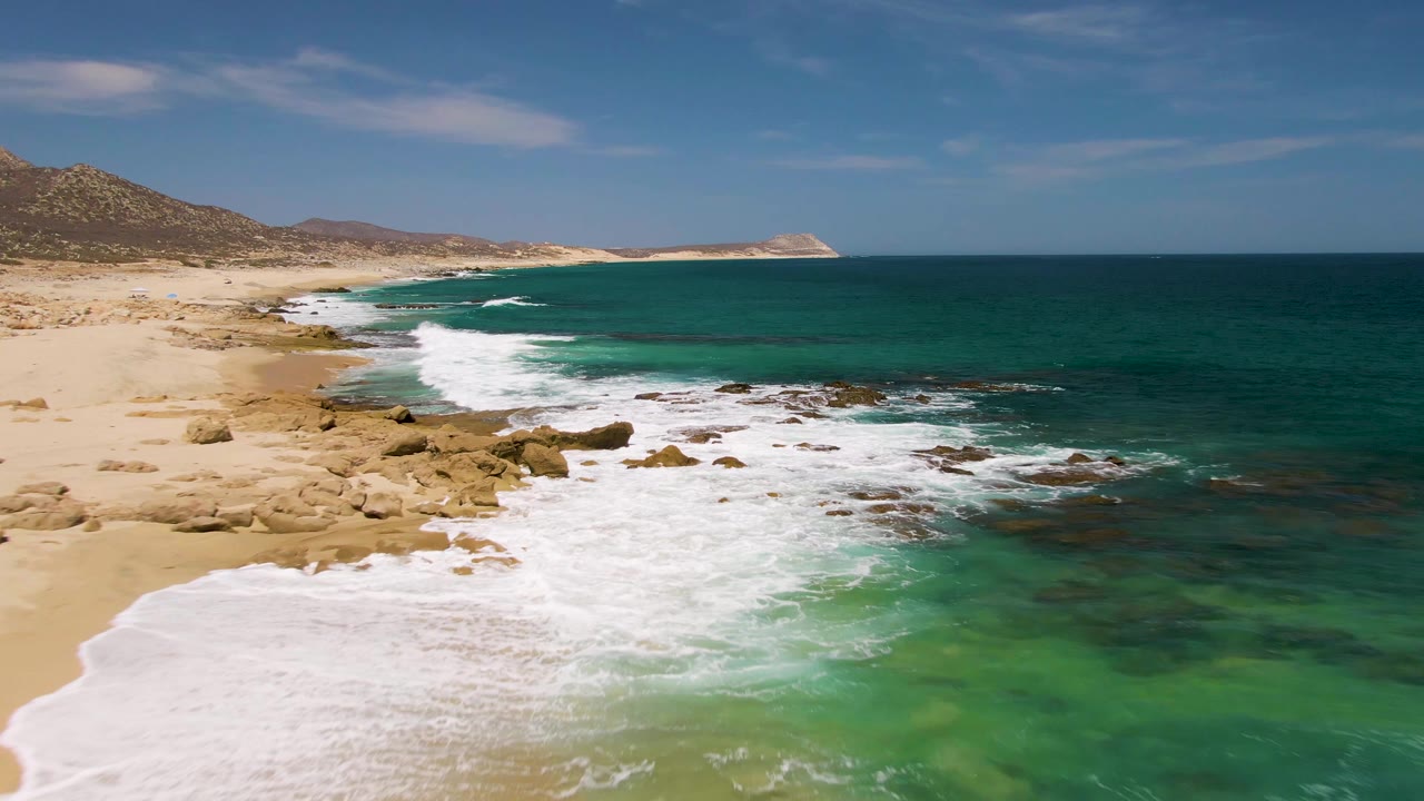 At Buzzards Beach in Baja California Sur, white foam pushes farther across the nearshore rocks while tan beach and low hills remain at left, with no visible people.
