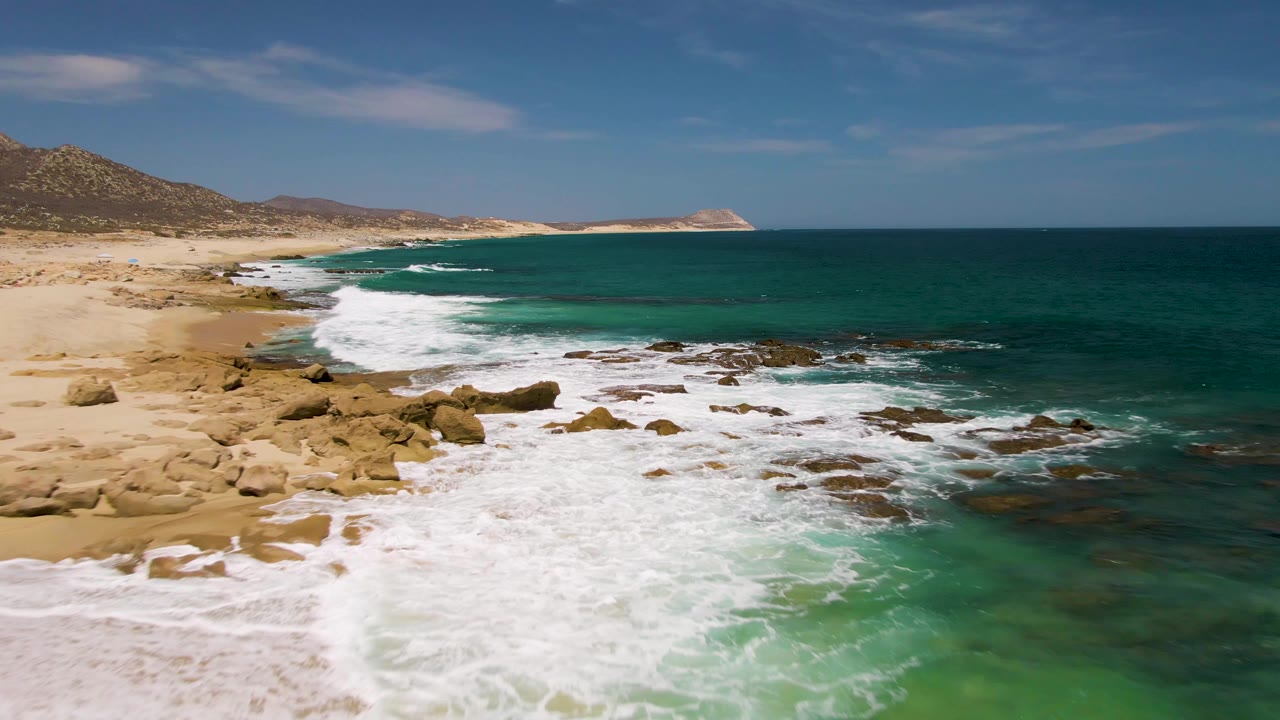 At Buzzards Beach in Baja California Sur, the surf thickens across the foreground rocks as turquoise shallows meet darker offshore water, with no visible people.