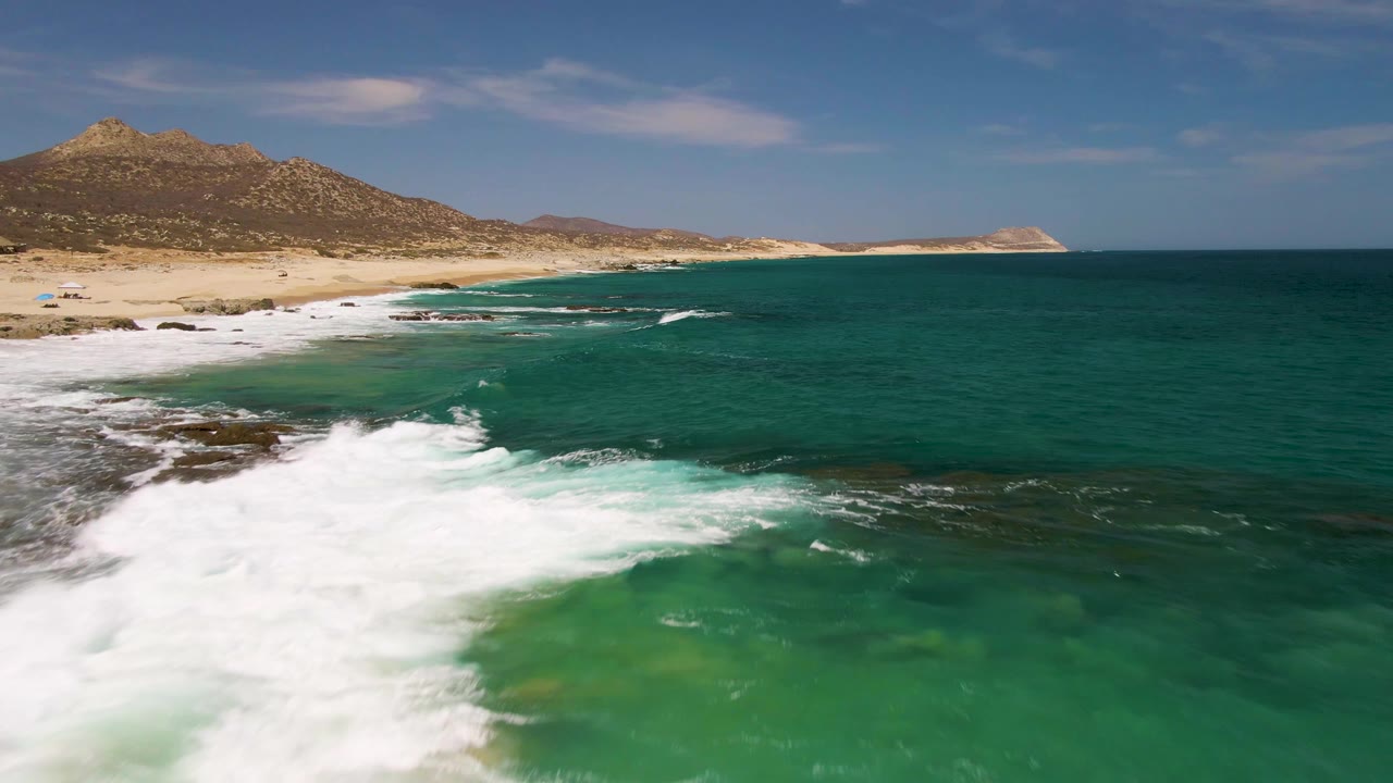 Aerial frame at Buzzards Beach in Baja California Sur with a larger sheet of white foam across the lower left, dark rocks near the shore, teal ocean to the right, and no visible people.