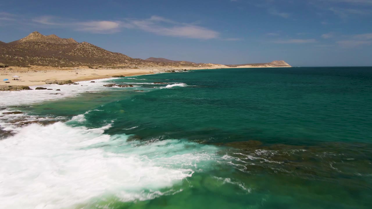 Aerial view at Buzzards Beach in Baja California Sur with foamy surf filling the lower left edge, clear green-blue water over darker submerged rock patches, sandy beach beyond, and no visible people.
