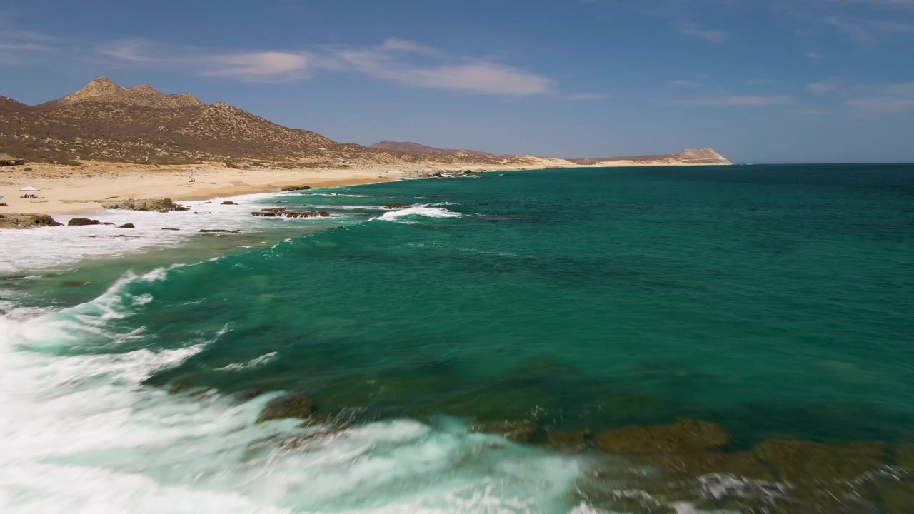 Aerial frame at Buzzards Beach in Baja California Sur with the coastline running left to right, white surf along the lower edge, deeper green water in the midground, distant hills, and no visible people.