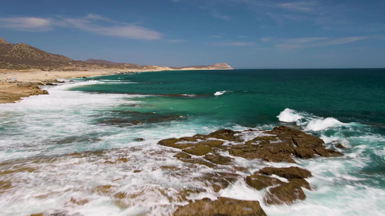 At Buzzards Beach in Baja California Sur, turquoise surf breaks across dark foreground rocks with a sandy beach, low hills, blue sky, and no visible people.
