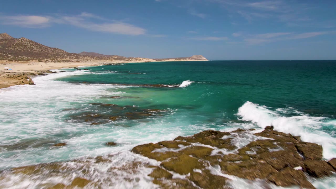 At Buzzards Beach in Baja California Sur, white foam spreads around foreground rocks while the sandy coast and low hills remain visible on the left, with no visible people.