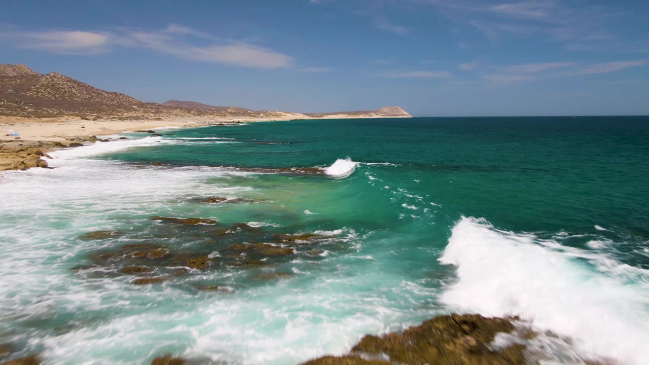 At Buzzards Beach in Baja California Sur, a larger white wave breaks at the right edge over shallow turquoise water, with reef rocks, beach, hills, and no visible people.