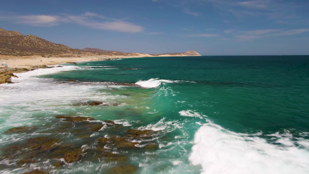 At Buzzards Beach in Baja California Sur, the shoreline opens wider with foamy water over rocks in the foreground, open turquoise ocean to the right, and no visible people.