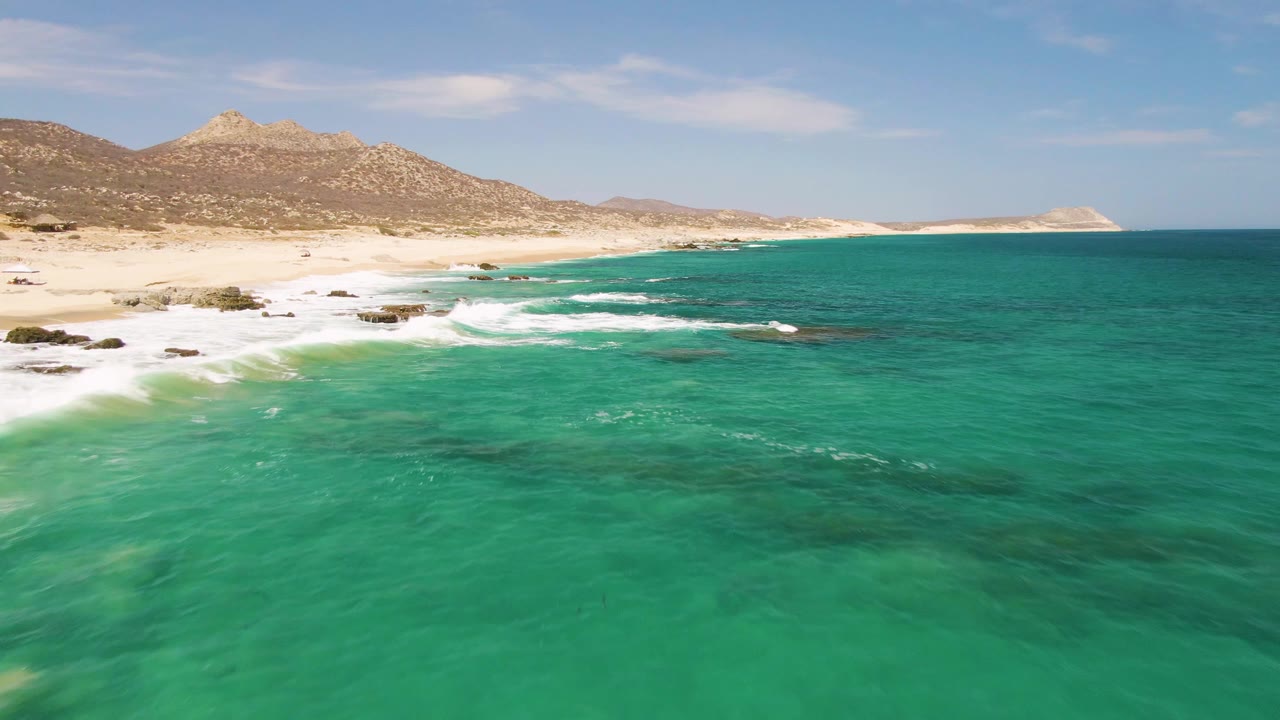 Aerial view over Buzzards Beach with clear turquoise water in the foreground, a sandy shoreline and rocky outcrops at left, tan hills in the distance, and no visible people.