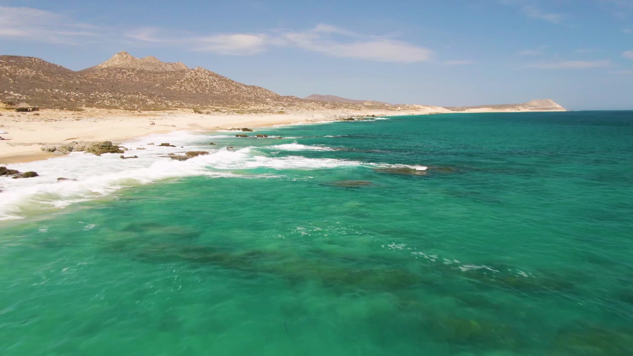 Buzzards Beach appears from above with transparent turquoise shallows across the foreground, a pale sandy curve at left, white wave lines near shore, and no visible people.