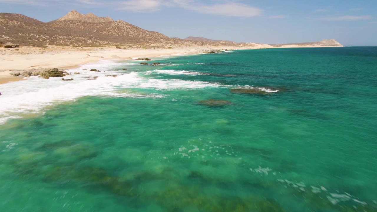 A higher coastal view of Buzzards Beach shows darker submerged patches in green-blue water, white surf touching the sand, rocky areas near shore, and no visible people.