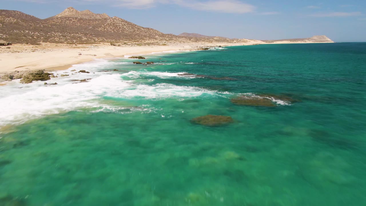 Waves breaking over rocks beside turquoise water and sandy shore