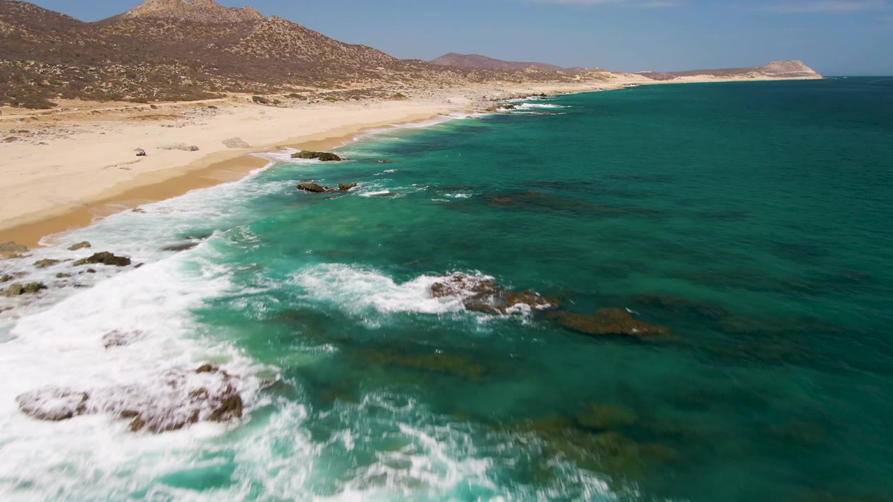 Aerial view at Buzzards Beach, Baja California Sur, with white surf hitting shoreline rocks, pale sand and dry hills in the background, and no visible people.
