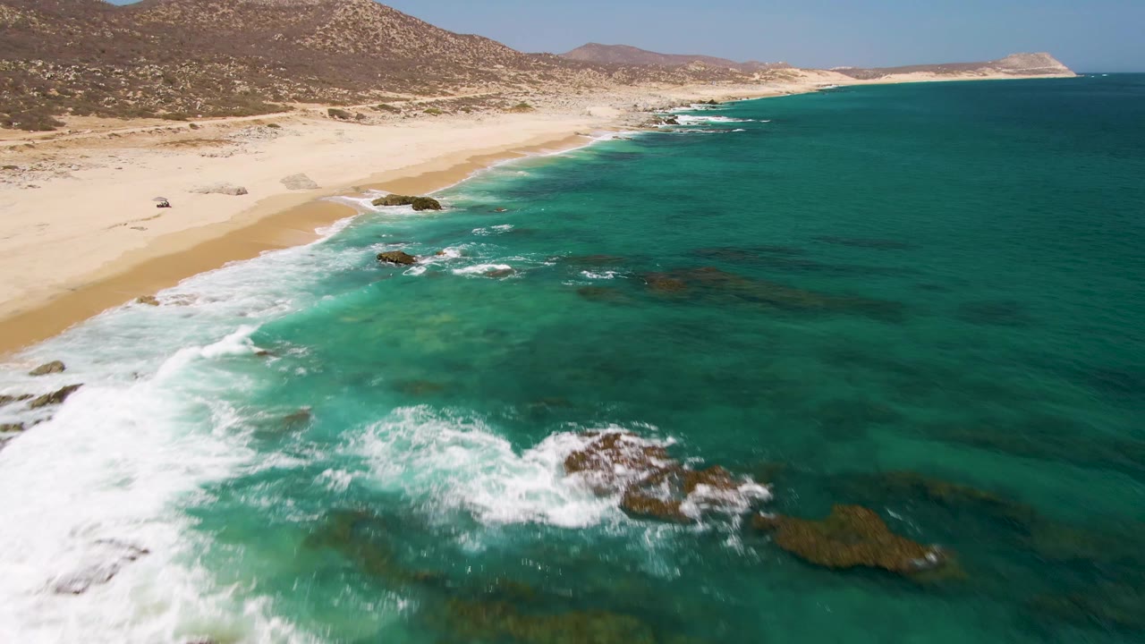 Drone-style aerial frame over Buzzards Beach showing foamy waves wrapping around dark rocks, shallow green-blue water, pale sand, and no visible people.