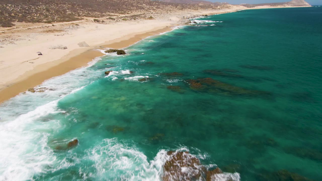 Aerial frame from Buzzards Beach with turquoise water filling most of the view, waves breaking along the sand, submerged rocks offshore, and no visible people.