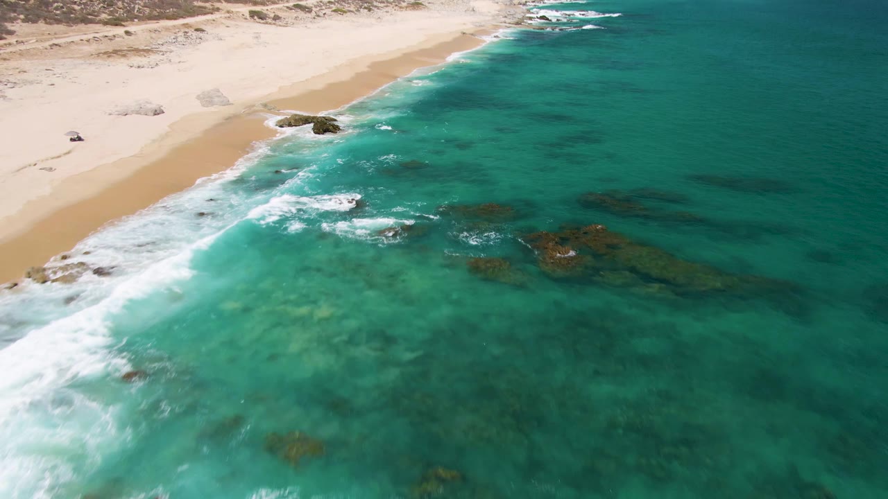 High aerial angle over shallow turquoise water, white surf, rocks, and empty sand.
