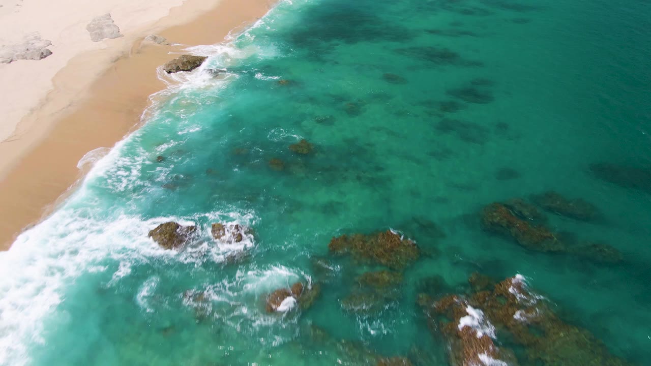 Aerial frame from Buzzards Beach, Baja California Sur, showing turquoise water, white surf around rocky outcrops, pale sand at upper left, and no visible people.