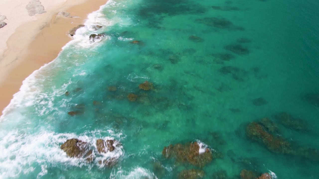 Aerial frame from Buzzards Beach, Baja California Sur, with surf spreading across shallow turquoise water, several dark rocks below the surface, sand along the left edge, and no visible people.