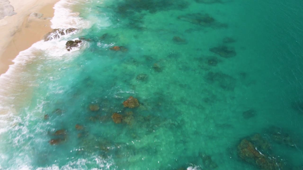 Aerial frame from Buzzards Beach, Baja California Sur, showing green-blue clear water over scattered submerged rocks, a strip of foamy surf at left, and no visible people.