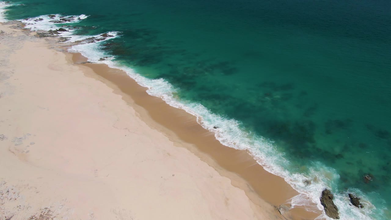 Overhead view of waves washing along pale sand and turquoise ocean.