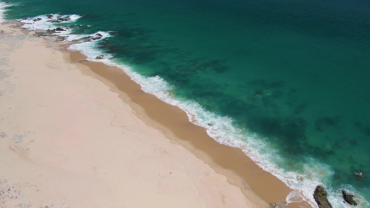 At 0:01, Buzzards Beach appears from above with a narrow band of wet sand, white foam advancing along the shore, darker underwater patches in the turquoise water, and no visible people.