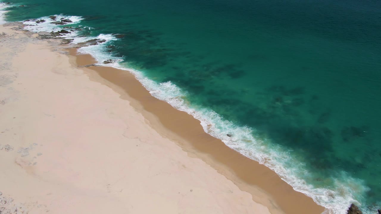 At 0:02, the overhead view shows Buzzards Beach with more open turquoise water across the upper right, pale sand below, a white surf edge, dark rocks near the shoreline, and no visible people.