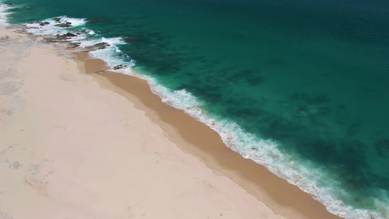 At 0:03, the aerial composition over Buzzards Beach keeps the pale empty sand broad on the left, green-blue water on the right, white foam crossing diagonally, faint beach marks, and no visible people.