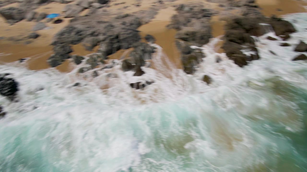 Overhead surf crashing around dark rocks on tan sand