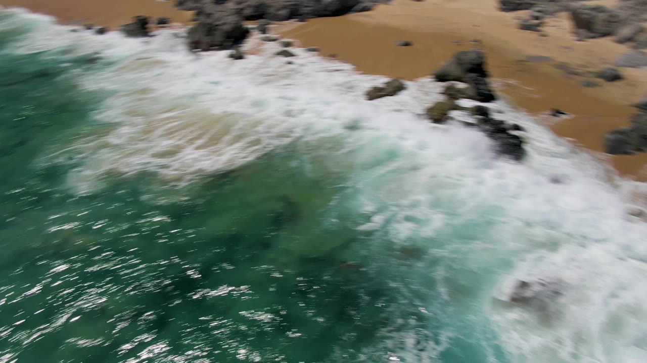 Aerial frame of white foam spreading over shoreline rocks and sand at Buzzards Beach, with deeper turquoise water filling the lower half and no visible people.