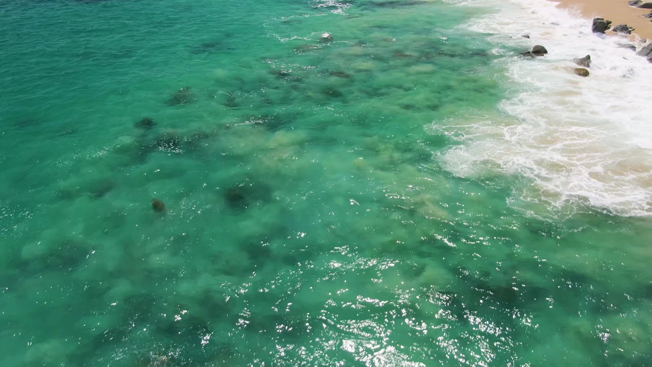 High aerial view of clear turquoise water at Buzzards Beach, with a white surf line along the sandy shore, scattered rocks, sun sparkle, and no visible people.