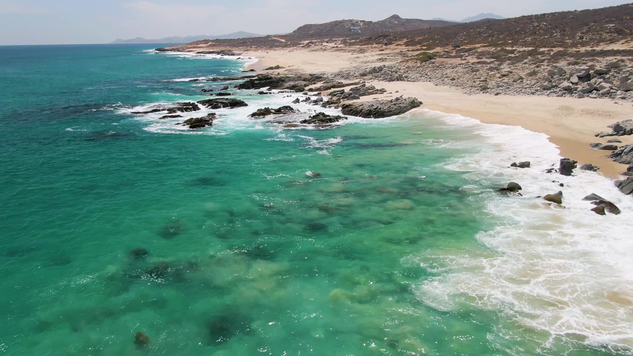 Wide aerial coastal view at Buzzards Beach showing turquoise ocean, white surf, dark rock clusters, tan sand, low hills in the background, and no visible people.