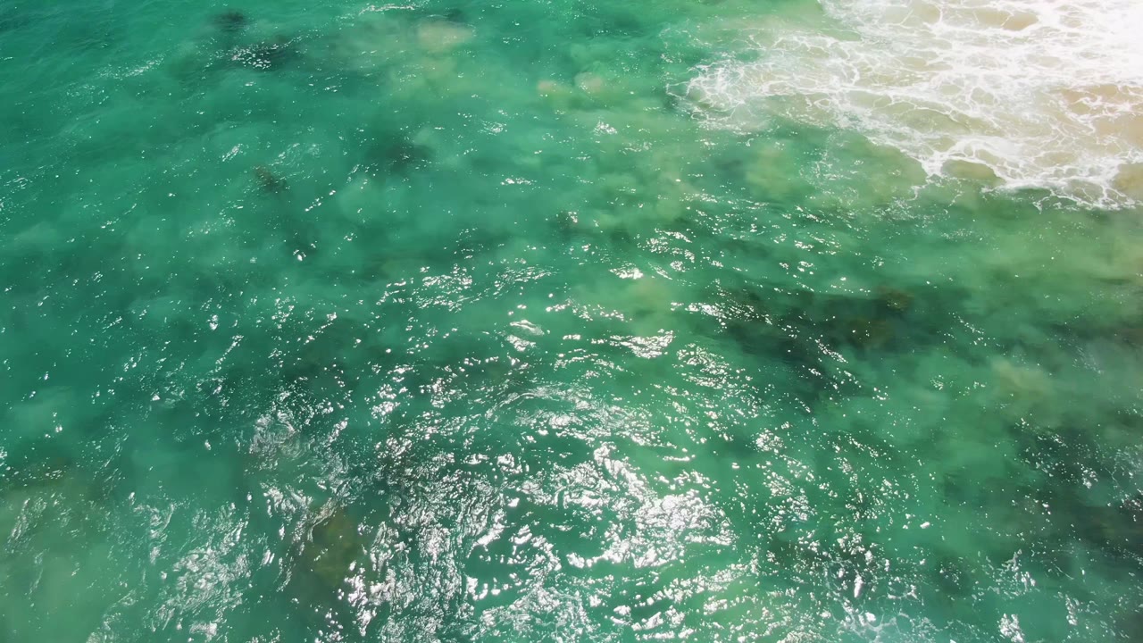 Aerial view over clear turquoise water at Buzzards Beach, Baja California Sur, with white surf entering from the upper edge and no visible people.