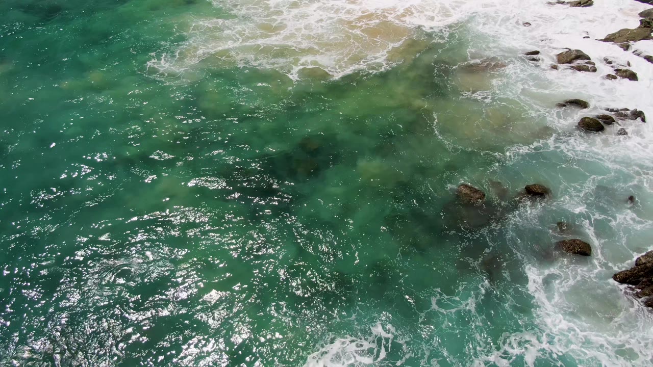 Clear green-blue water beside breaking foam and scattered rocks