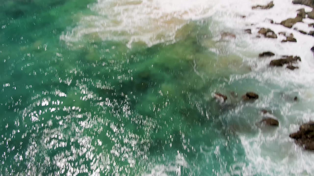 Aerial view at Buzzards Beach, Baja California Sur, with bright reflections on turquoise water, white surf at the right side, exposed rocks, and no visible people.
