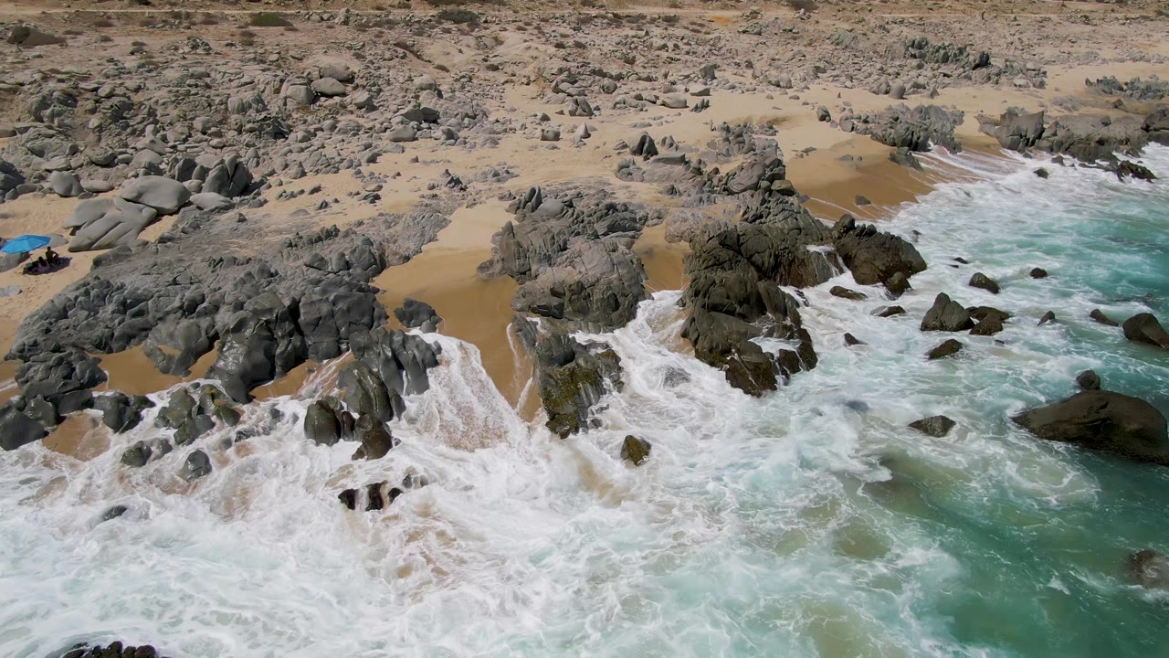 Aerial frame over Buzzards Beach in Baja California Sur showing white surf washing across dark rocks, tan sand, turquoise water, and no visible people.